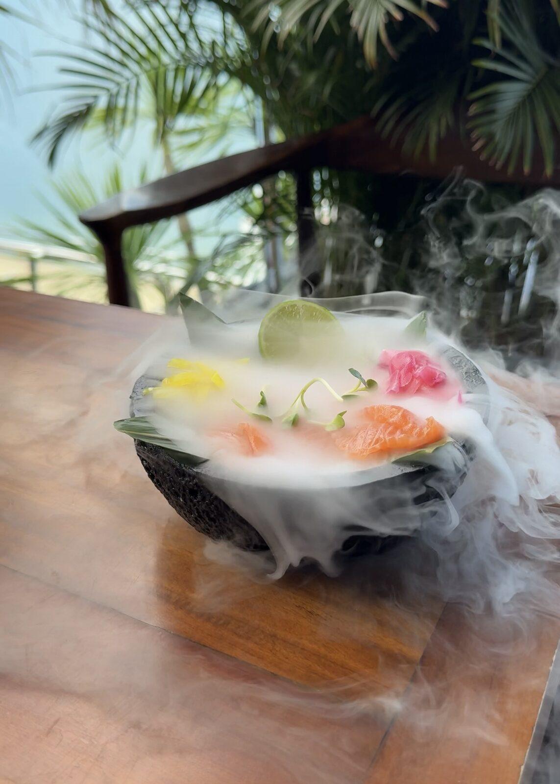 Salmon sashimi in a stone bowl with smoke, palm trees behind